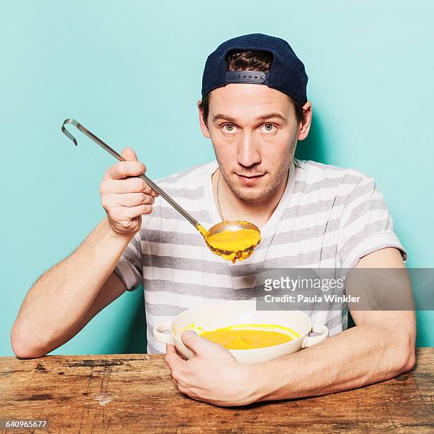 portrait of confident man having pumpkin soup at table against blue background - soup on spoon stock pictures, royalty-free photos & images