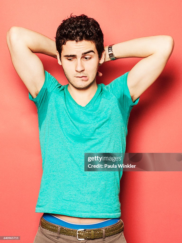 Young man biting lip while looking at sweaty armpit against red background