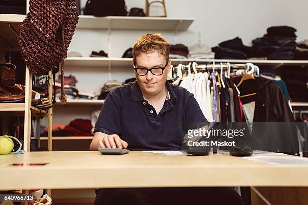 young man calculating while sitting at checkout counter in clothing store - disability collection stock pictures, royalty-free photos & images