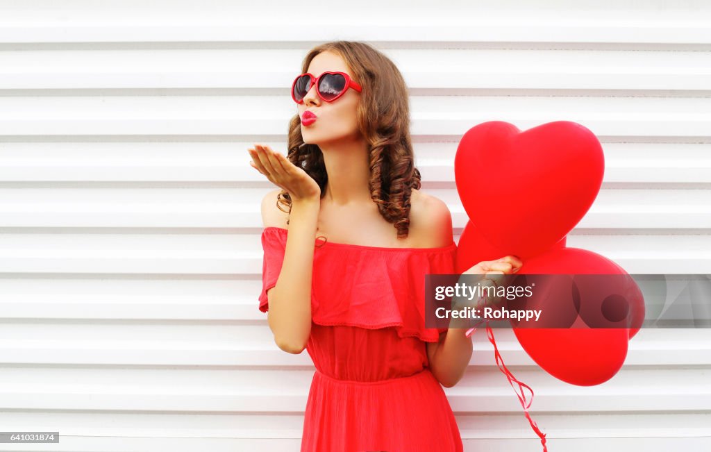 Portrait woman in red dress sends air kiss with balloon heart shape over white background