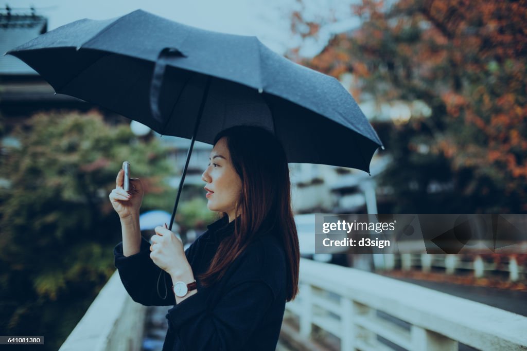 Young female traveller holding umbrella and taking pictures with smartphone on street