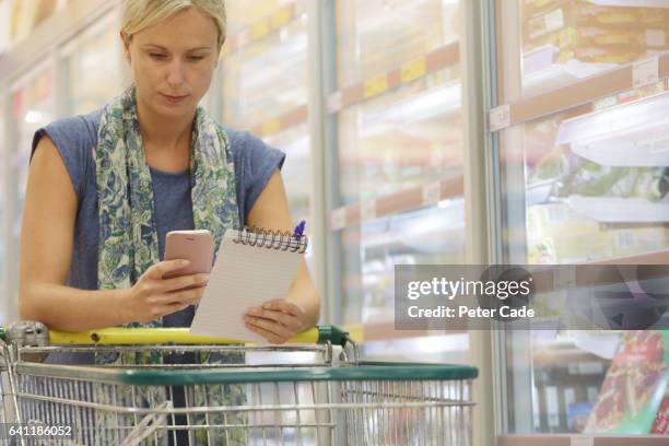 woman with shopping trolley looking at phone and list - supermarket trolley female stock-fotos und bilder