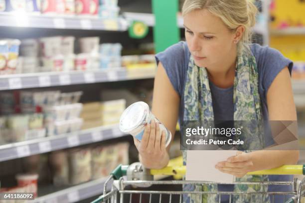 woman holing shopping list looking at information on yogurt pot - grocery food stockfoto's en -beelden