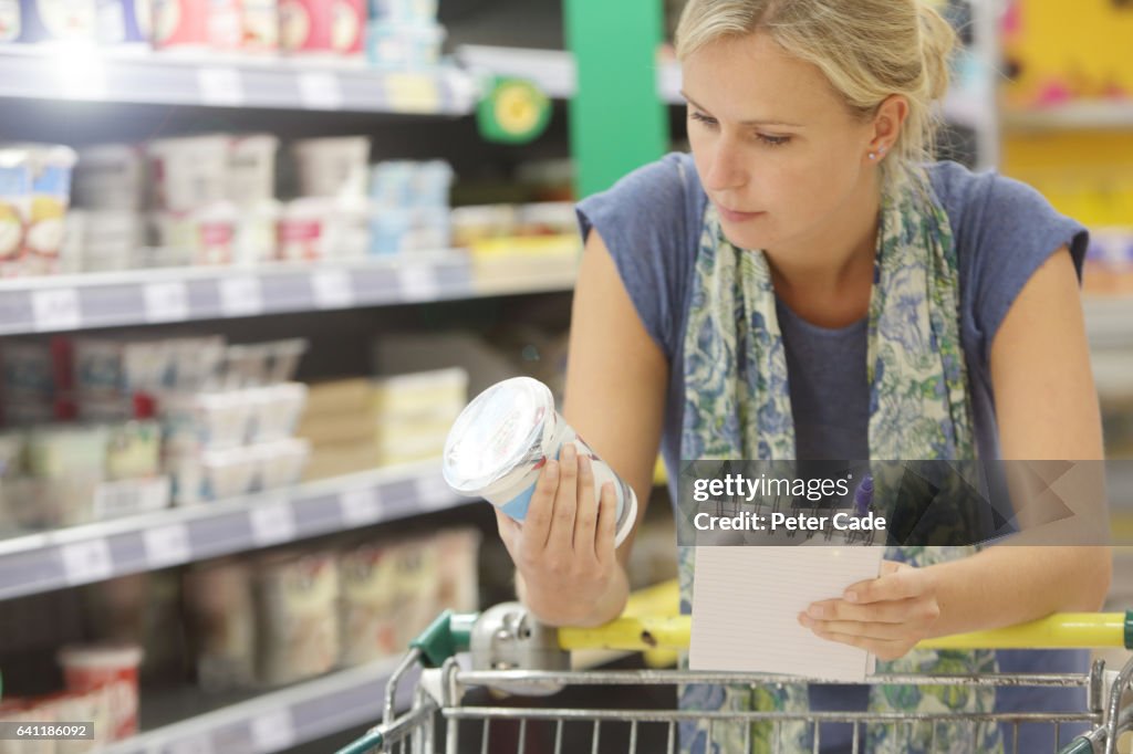 Woman holing shopping list looking at information on yogurt pot