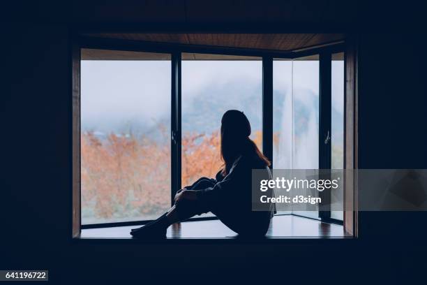 woman sitting alone on windowsill looking through window in autumn - back lit home stock pictures, royalty-free photos & images