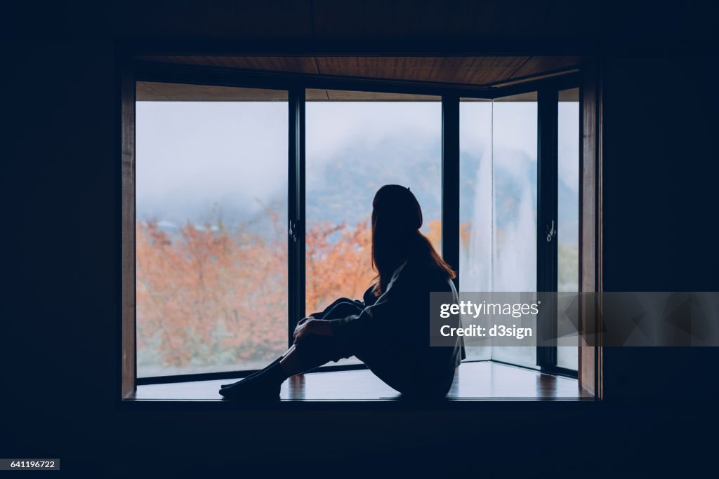 Woman sitting alone on windowsill looking through window in Autumn