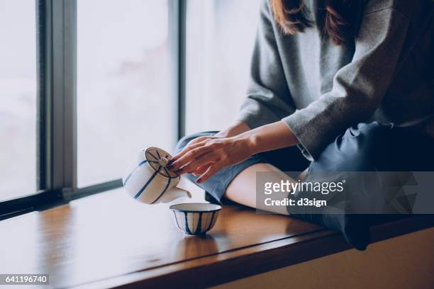 woman sitting on the windowsill and pouring a cup of tea - feierliche veranstaltung stock-fotos und bilder