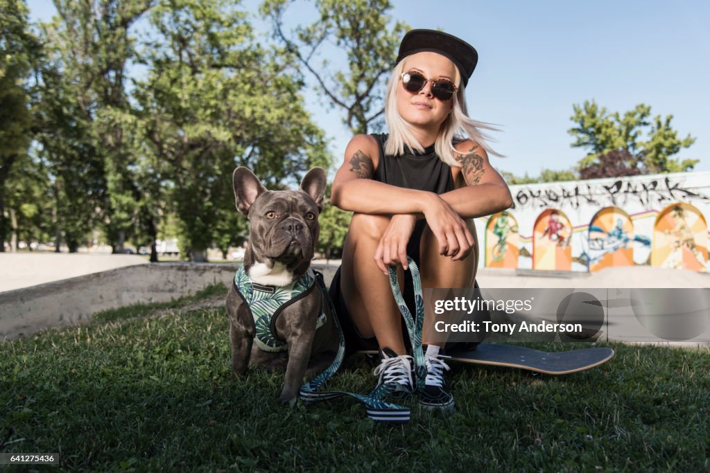 Young woman sitting on skateboard at skatepark with her dog