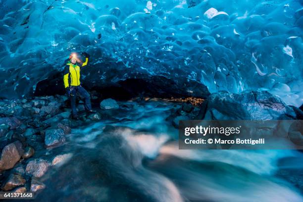 man with yellow coat by a flowing river inside a crystal ice cave in winter. iceland. - crystal caves stock-fotos und bilder
