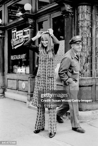 Portrait of an unidentified model, dressed in a matching brocade trouser suit and hat, as she poses in front of PJ Clarke's saloon , New York, New...