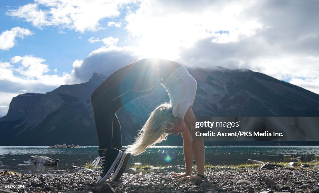 Young woman performs back arch on shore of mountain lake