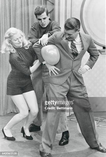 Bud Collyer, the show's "stunt master," offers his services as Doris Haley and Jim Holland try to force a balloon past his elbow without its...