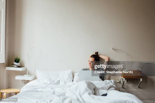 women in bed looking out window and smiling with laptop - comfortable bedroom stock pictures, royalty-free photos & images