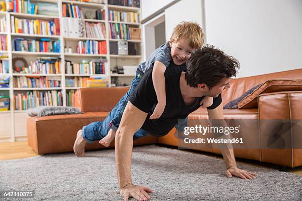 father doing push ups in living room with son on his back - flexiones fotografías e imágenes de stock