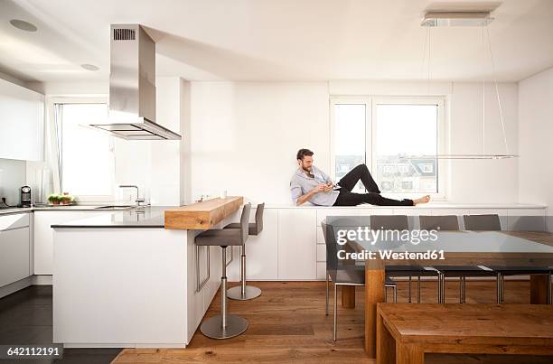 man lying on sideboard in his open plan kitchen using smartphone - acostado de lado fotografías e imágenes de stock