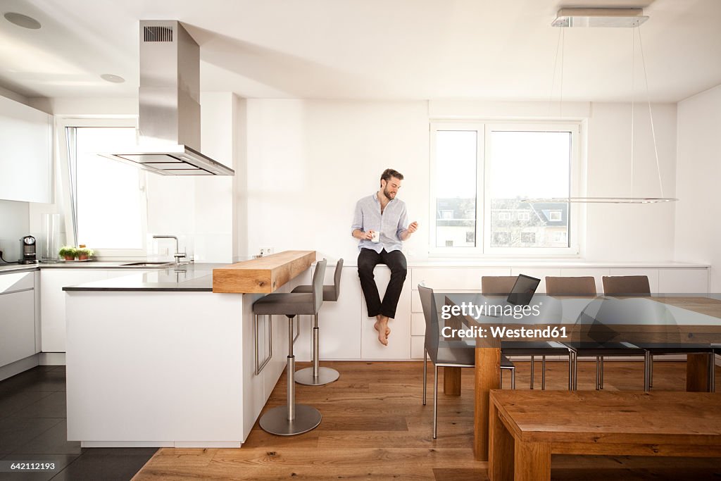 Man with coffee cup sitting on sideboard in his open plan kitchen looking at smartphone