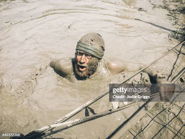 man covered in mud emerging from a muddy pool of water during a challenge event - mud run stock-fotos und bilder