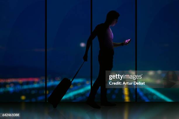 stockillustraties, clipart, cartoons en iconen met silhouette of a guy checking his smartphone ready to departure on a airport at night with bokeh lights during a travel with trolley suitcase using application technology to get information about the trip. - looking