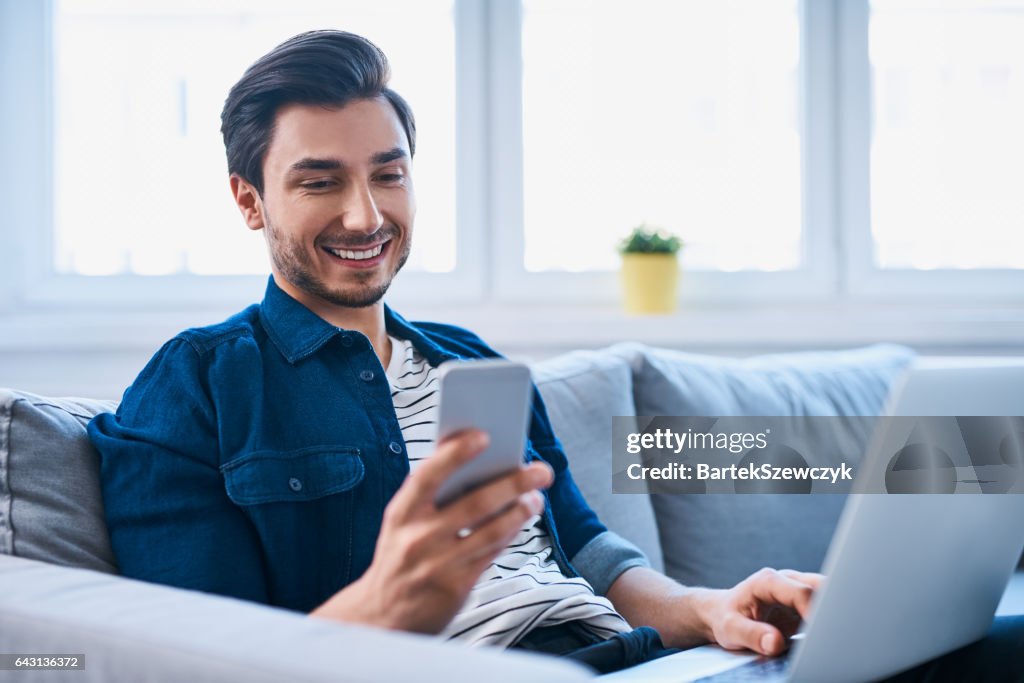 Junger Mann sitzt entspannt auf Sofa mit Laptop und smartphone