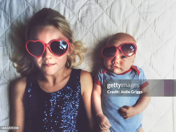 young girl and baby laying beside each other wearing love heart glasses - sisters stock-fotos und bilder