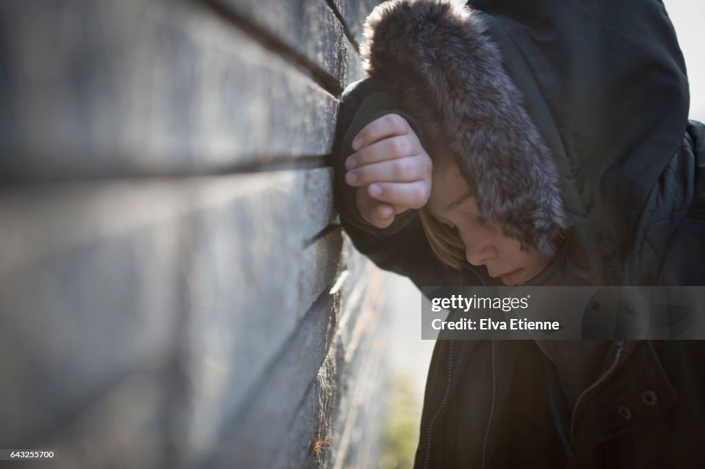 Teenage boy in hooded coat, leaning against a wall in despair
