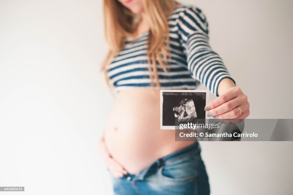 Pregnant caucasian woman wearing a striped shirt and blue jeans. Holding an ultrasound pic of the baby in her tummy. Nude belly.