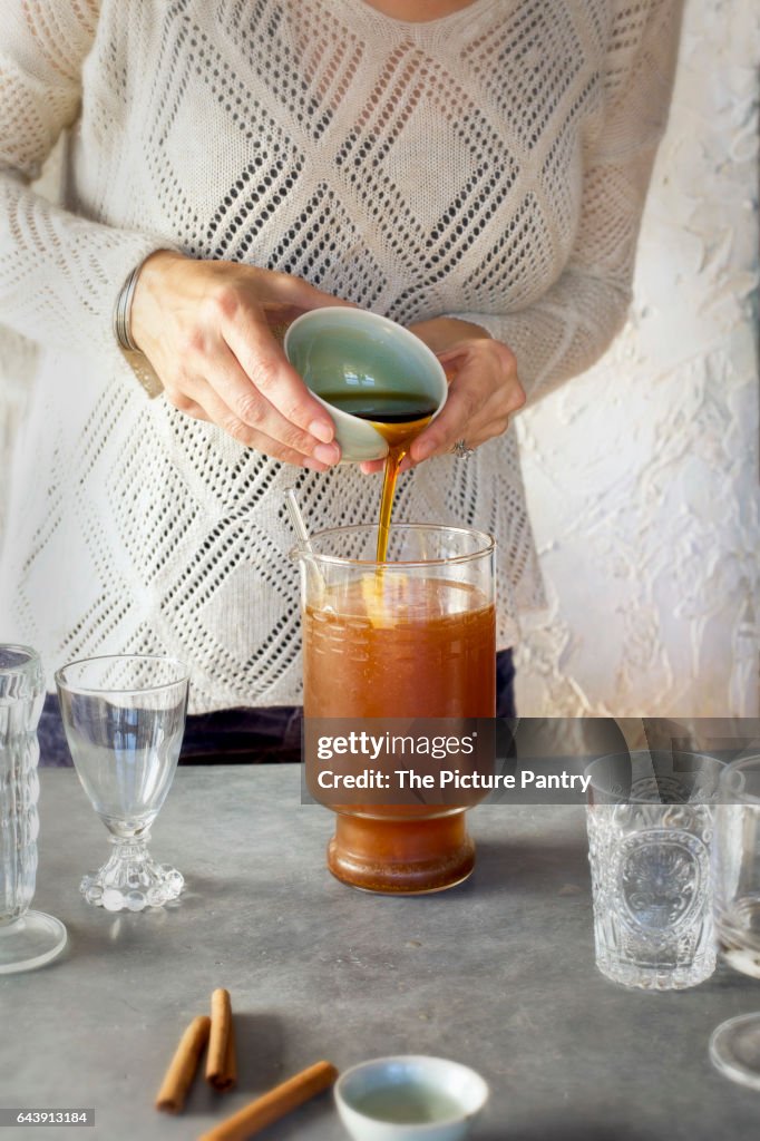 A Women is pouring maple syrup into Almond Lime Italian Soda in glassware. Photographed on dark gray background.