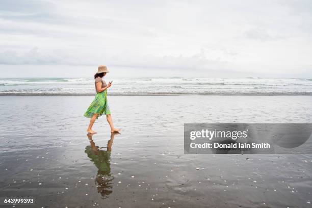mixed race woman walking on beach with cell phone - new zealand beach stock pictures, royalty-free photos & images