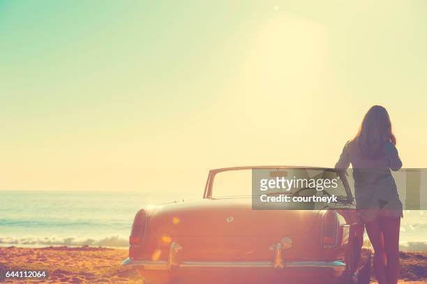 woman leaning on her car at the beach. car is a classic convertible. she is looing out at the ocean at sunrise or sunset. looking very relaxed on vacation. - road trip new south wales stock pictures, royalty-free photos & images