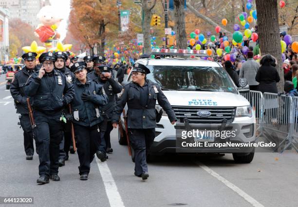 New York Police Department officers with riot gear walk past a police cruiser, with colorful balloons visible in the background, during the 90th...