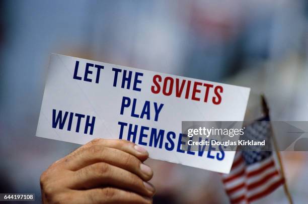 Person attending the 1984 Summer Olympic Games in Los Angeles holds a bumper sticker which reads, "Let the Soviets Play with Themselves," referring...