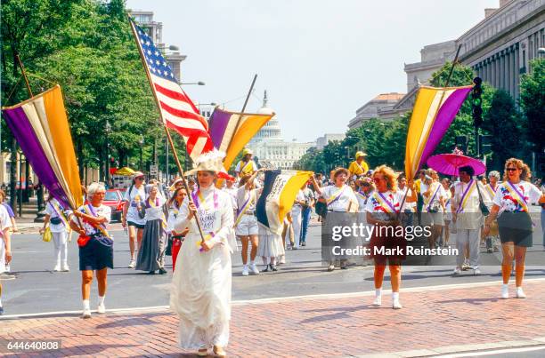 View of marchers, among them one dressed as Carrie Chapman Catt who carries an American flag, as they walk along Pennsylvania Avenue during a rally...