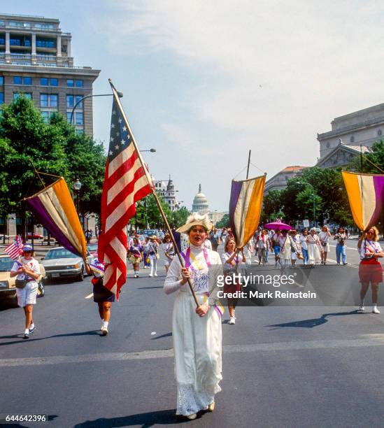 View of marchers, among them one dressed as suffragist Carrie Chapman Catt who carries an American flag, as they walk along Pennsylvania Avenue...