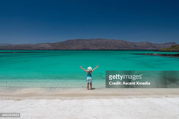 woman stands on sandy beach, lifts arms overhead - océano pacífico fotografías e imágenes de stock