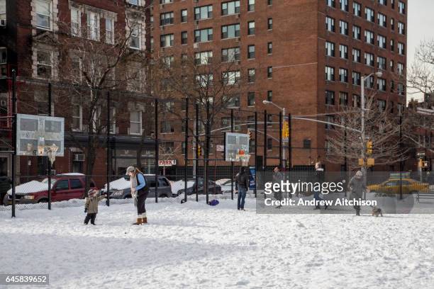 Woman plays with her young child with the snow, behind are three men playing basketball as a woman walks past with her dog just off Husdon Street,...