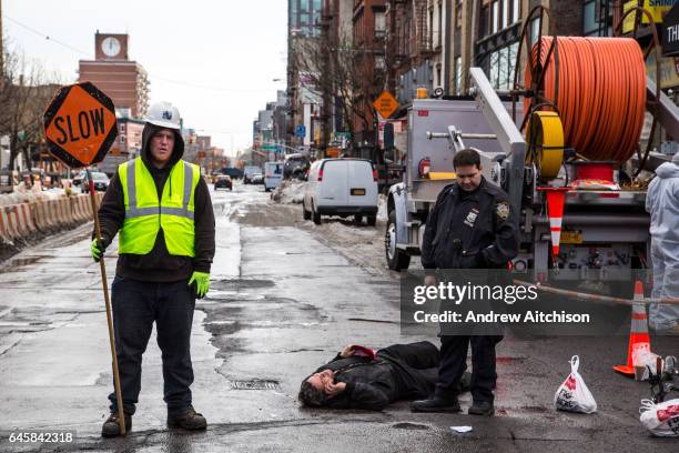 Man lies in the street talking on his phone after falling off his bike, an NYPD police officer stands next to him and workman holds a SLOW sign to...