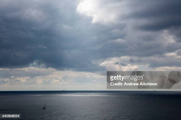 Sail boat sails across the tranquil waters of The English Chanel in the forefront, and two large shipping containers sail along the horizon line as...