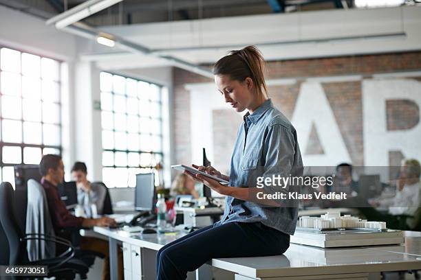 woman sitting on desk with tablet at, open office - vista lateral - fotografias e filmes do acervo