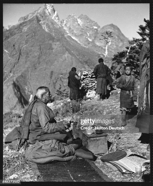 Lamaist priest chants prayers and repeats passages from religious scrolls while his helpers make a pile of faggots for a funeral pyre.