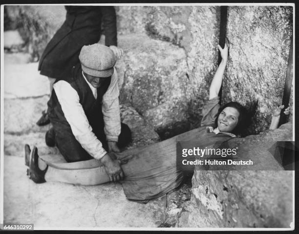 Man holds a girl's legs as she prepares to kiss the Blarney Stone. The stone, a block below the castle battlements, must be reached by leaning over...