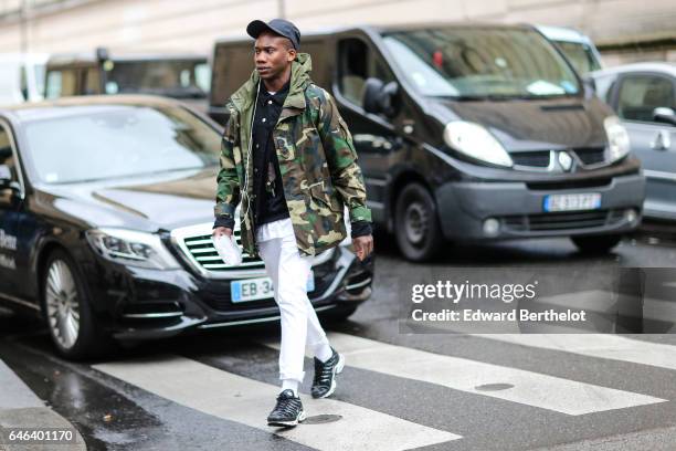 Guest wears a cap, a military camouflage print coat, white pants, and sneakers shoes, outside the JOUR/NE show, during Paris Fashion Week Womenswear...
