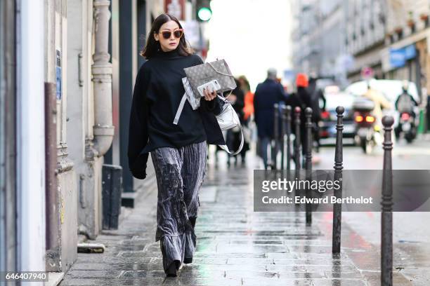Guest wears sunglasses, a black top with long sleeves, gray large pants, and black shoes with large heels, outside the AALTO show, during Paris...