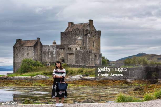 Asian tourist taking selfie with smartphone camera on selfie stick in front of Eilean Donan Castle in Loch Duich, Ross and Cromarty, Western...