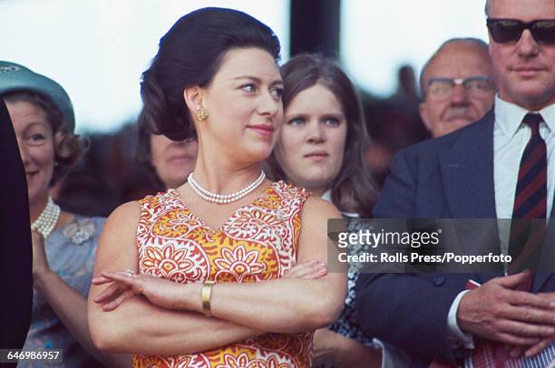 Princess Margaret, Countess of Snowdon pictured wearing a pearl necklace and orange, white and yellow patterned summer dress as she watches the...