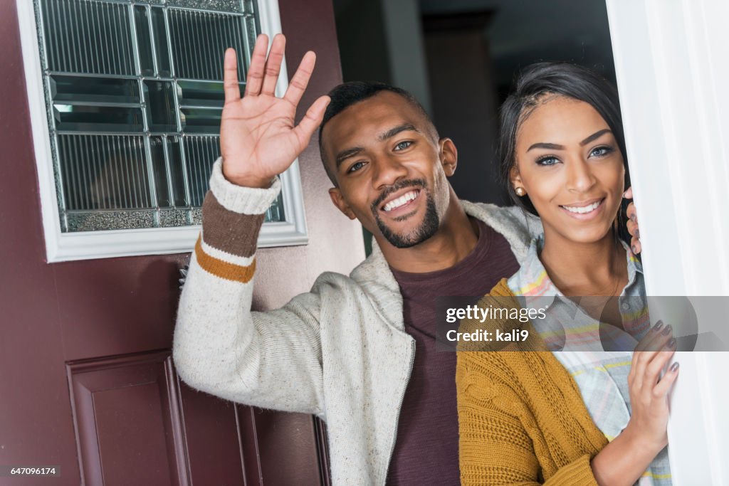 Young mixed race couple standing in front doorway waving