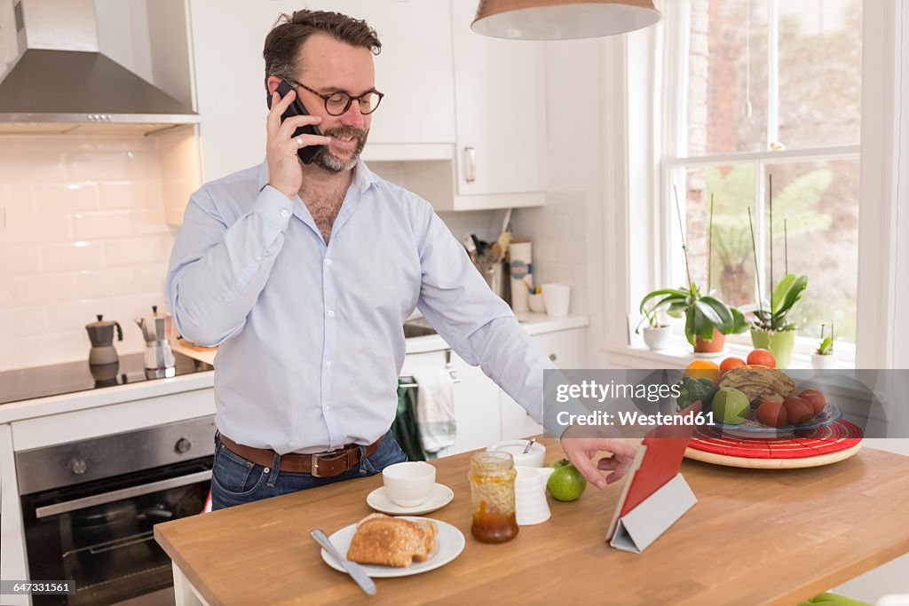 Man standing at breakfast table in the kitchen telephoning with smartphone while using digital tablet