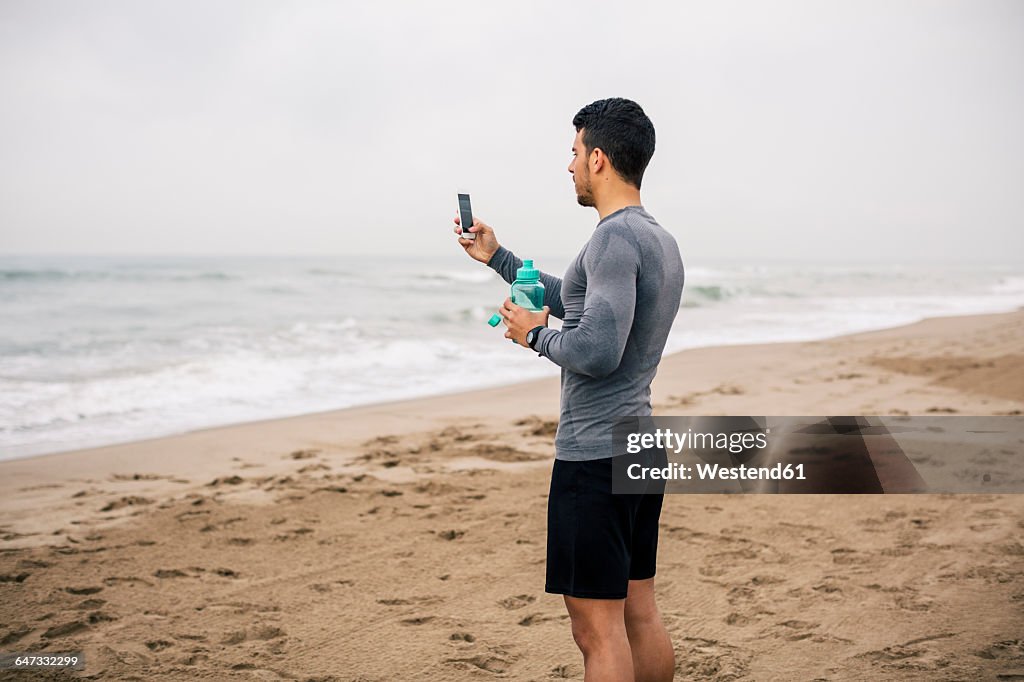 Sportive young man with cell phone and drinking bottle on the beach