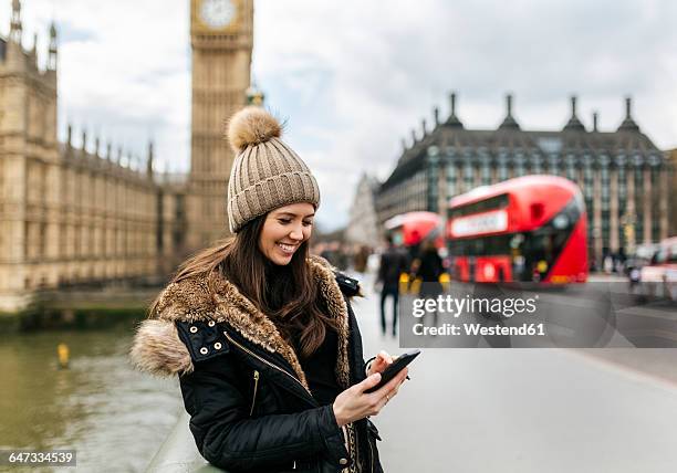 uk, london, smiling young woman looking at her smartphone in front of palace of westminster - bobble hat stock pictures, royalty-free photos & images