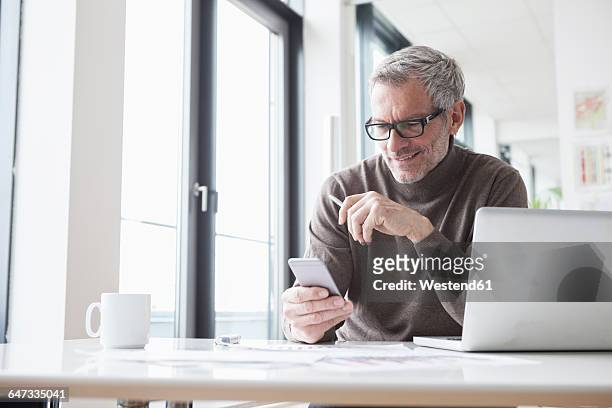 mature man sitting in office using laptop - business man looking at smart phone stock-fotos und bilder