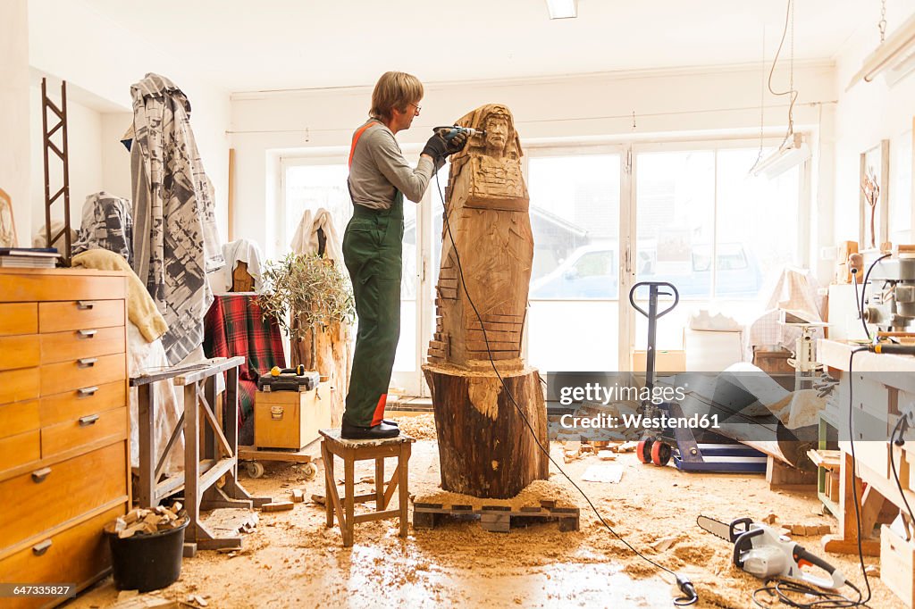 Wood carver in workshop working on sculpture with milling machine, standing on stool